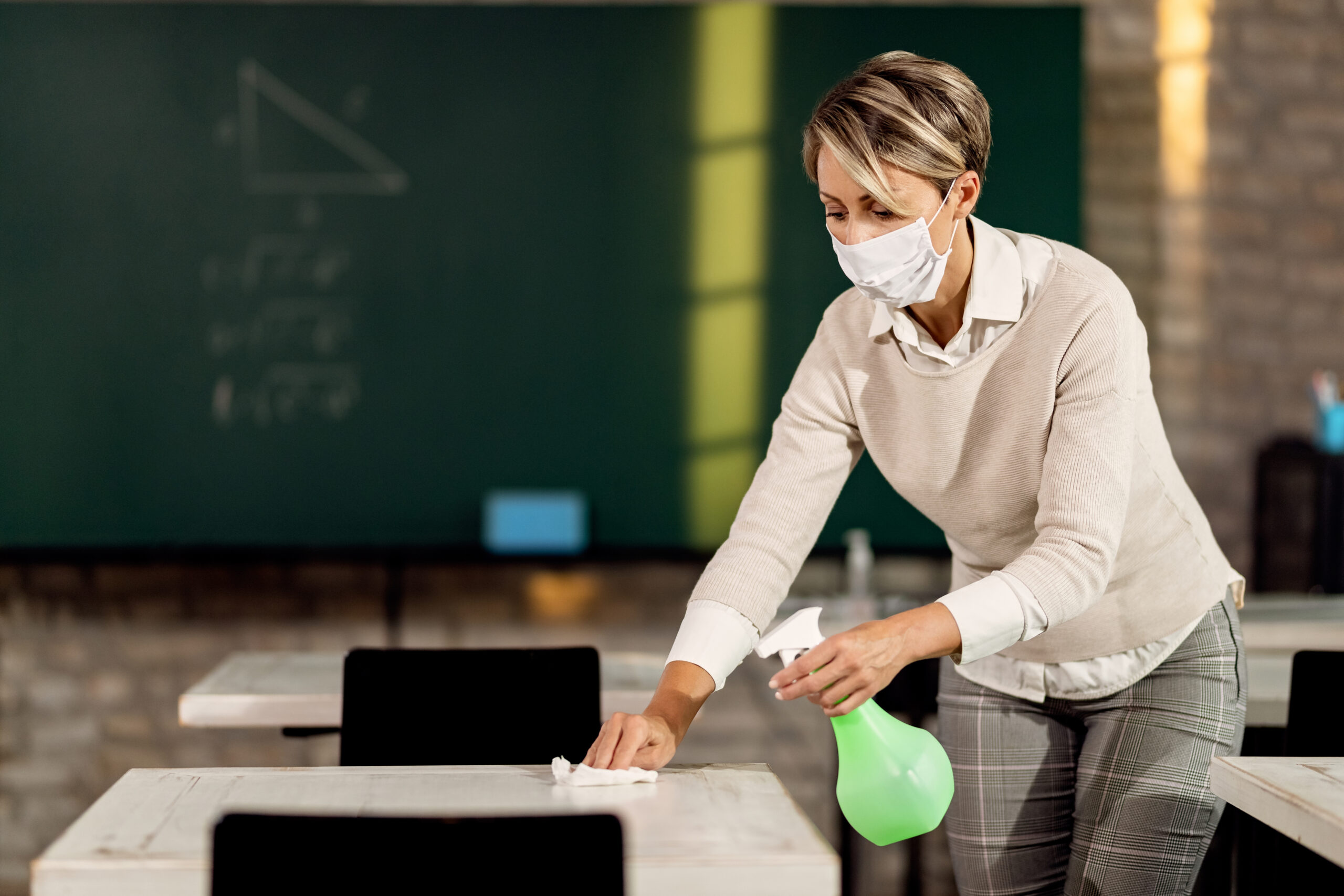 Teacher with face mask preparing classroom for kids and cleaning tables with disinfectant.